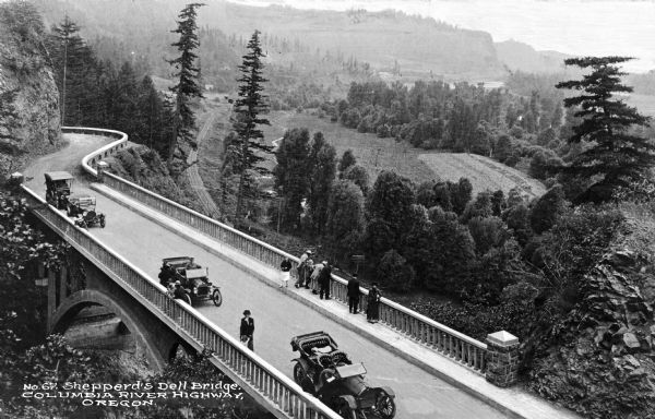 Elevated view of Shepperd's Dell Bridge on the Columbia River Highway, a historic scenic road. People stand near the railing to look out over the bridge. Railroad tracks run along below the bridge towards hills in the distance. A rock formation borders the road on the left and trees and forested hills are in the background. Caption reads: "Shepperd's Dell Bridge. Columbia River Highway, Oregon."