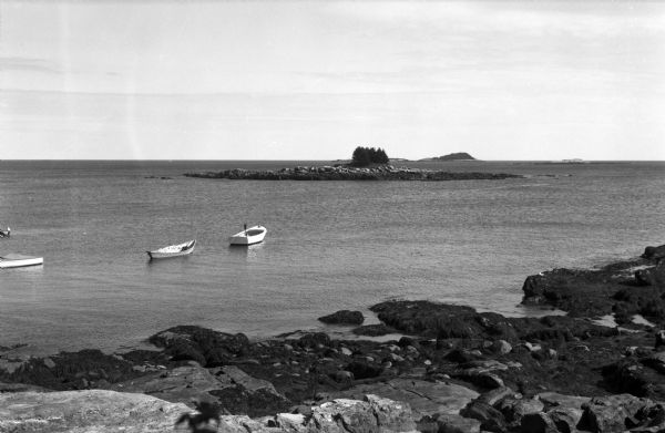 Elevated view of three rowboats floating in the water off a rocky shore, and in the distance several rocky islands with trees.