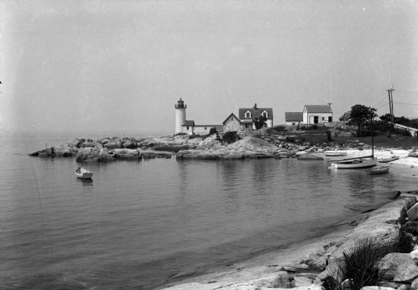 View along shoreline toward a lighthouse and homes standing on Rocky Point. There are several boats anchored at the shore and in the water.