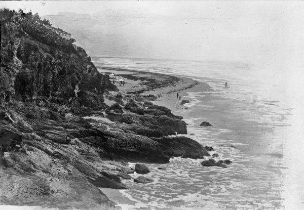 Elevated view toward a dwelling that overlooks a steep rocky shoreline over the ocean below. Several people walk along the sand.