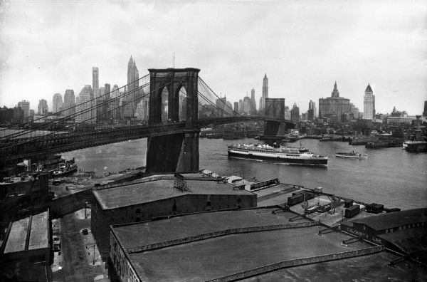 View of the East River and the Brooklyn Bridge. Several boats and ships sail down the river and industrial buildings and skyscrapers stand in the foreground and extend toward the horizon.