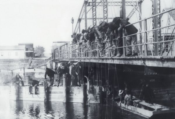 A large group of men are snagging sturgeon from a bridge over the Wolf River. A sturgeon is hanging from a line. More men are sitting on a bridge support, note the sign that says "NO PIPE SMOKING" with a man smoking a pipe. There is a boat under the bridge with 5 men in it. The fish were snagged for their eggs which were sold to the big city market and the fish discarded like cord wood to rot. The bridge was replaced between 1880 and 1890.