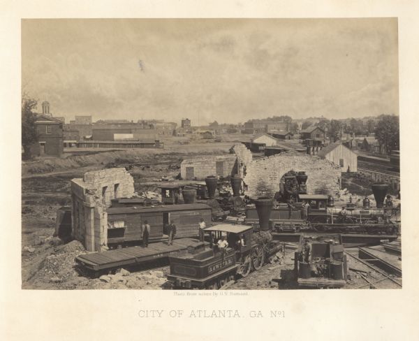 Elevated view of the ruins of the railroad roundhouse, with the city in the background. Men are posing on the train cars and engines.<br>Plate 45</br>