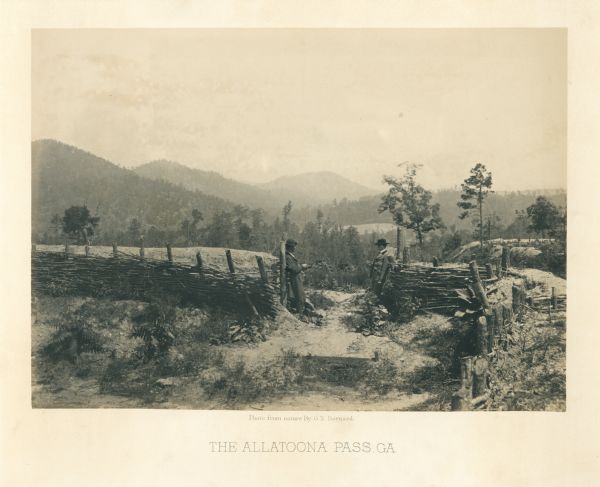 Two men stand on either side of an opening in the earthworks at Allatoona Pass. Mountains are in the distance.<br>Plate 28</br>
