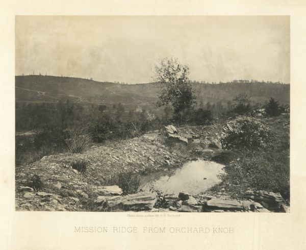 A rocky ridge with bushes and scrub atop Mission Ridge is in the foreground. Orchard Knob is in the distance. The valley where the fighting took place is in the middle.<br>Plate 09</br>