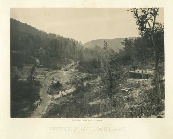 View down to valley of Union soldiers posing on the roof of a log building. A log bridge crosses a stream, and many trees are on the hillsides.<br>Plate 05</br>