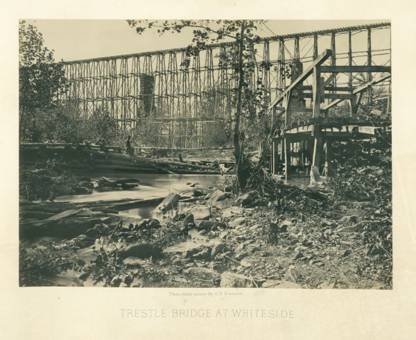 View from shoreline of tall trestle bridge on the railroad line crossing the river at Whiteside. Two men sit on logs in the river on the left. More men sit on a timber structure along the river on the right.<br>Plate 04</br>