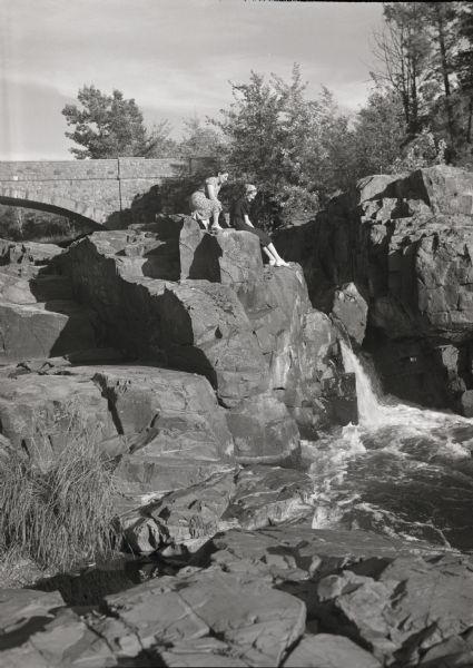Two women sitting on top of rocks in the Dells of the Eau Claire River. There is a bridge in the background on the left.