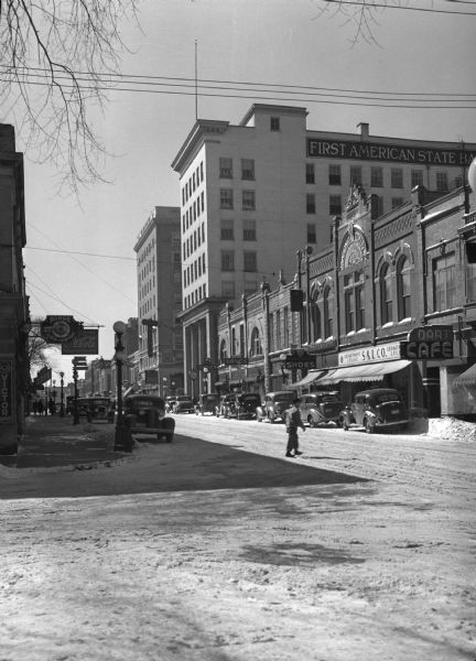 View from street of commercial area with storefronts and pedestrians. Many cars are parked on both sides of the street. Visible storefront signs read, "S & L Co. Department Store," "Dart Cafe," "Shoes," "Drugs," "Hotel ?," "Savoy Cafe," "Norge ? Refrigeration," and "First American State Bank."