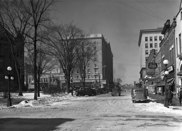 View of Scott Street looking west. There are automobiles parked on each side of the snowy street, and two men are standing on the corner holding shovels. Storefronts with signs are also visible, including "Damon Bros. Insurance," "Livingston's," "Cut Rate System Drugs Walgreen," and "Hotel Wausau."