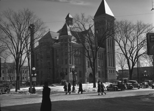 View from street of the Marathon County Courthouse, "Bet 3rd and 4th, Jefferson and Scott Streets". Men and women are crossing the snowy streets near parked automobiles.