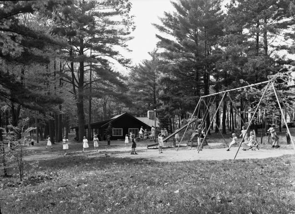 A view of children playing on the swingset, slide, and other playground equipment at a park. A wooden building is located behind the playground, and the park itself is located in the Wausau vicinity.