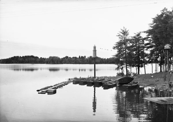 View of Oneida Colony, a northern lake resort. Small boats are lined up along the shore, and birds are gathered near a large birdhouse. A man is lifting one of the boats.