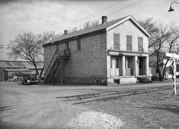 The former Astor (American Fur Trading Company) post building, now a museum of local history and fur trading history. There is a sign reading: "The Riverside Repairs Shop" over the entrance to the building, and railroad tracks are in the foreground.