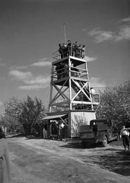 View across road of an observation tower overlooking extensive apple orchards, a tourist attraction in season. Many people are standing below the tower where a snack shop is located beneath an awning. Many people are crowded on the stairs waiting to get to the top of the tower. A truck is also parked nearby.