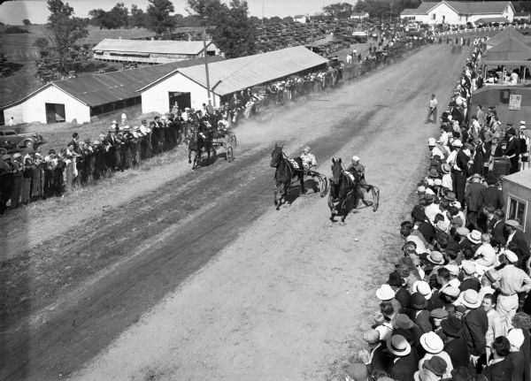 Elevated view of four men and their horses harness racing down a road, with crowds of spectators on the sides, during a county fair in southwestern Wisconsin.