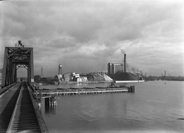 View from railroad bridge of the harbor at Green Bay. On the other side of the harbor, there are many industrial buildings, including Northern Paper Mills, and a water tower. A man is walking on the bridge on the railroad tracks.