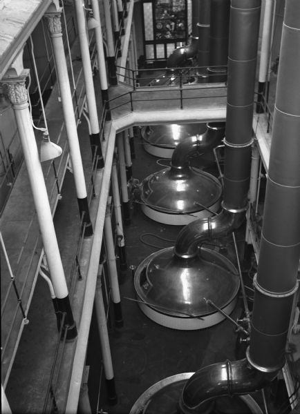 View from above of brewing kettles in the Pabst brewery in Milwaukee. There are workers standing on the ground floor, and in the far background is a large stained glass window.