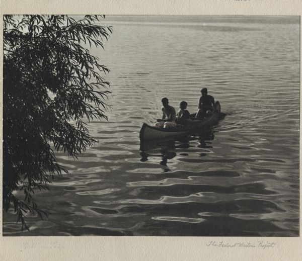 View from shoreline of four individuals in a canoe on Lake Mendota, framed with branches from a tree on the left side of the image.