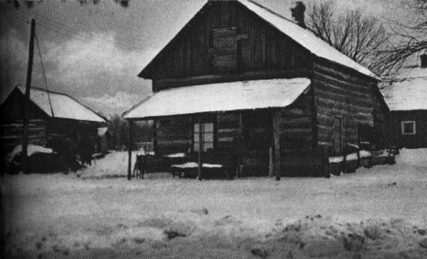 A view of a "log cabin of a pioneer". Two other log cabins are visible in the background.