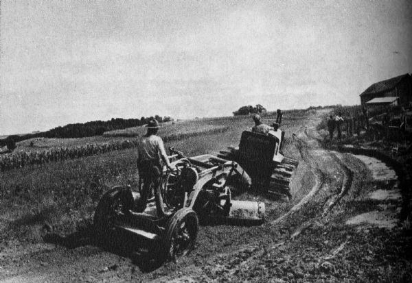 A view of two men on tractors, contour plowing a field. Two other men stand near a dirt road.