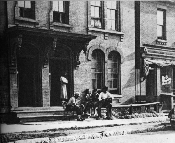 Five people sitting on a sidewalk outside of a building. A woman stands on a stoop in the doorway.