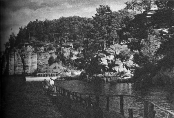 A view of the Dells of the Wisconsin River, with two people walking down a long dock/walkway leading to a boat.