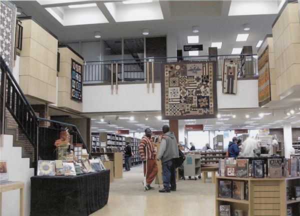 Interior view of La Crosse library. Two men are talking near a display of books. A balcony in the background has a display of quilts.
