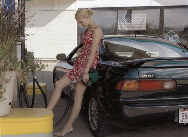 A blond-haired woman in a dress pumps gas.