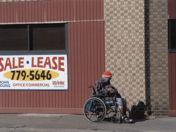 A man in a wheelchair is sitting outside a building with a "Sale - Lease" sign.