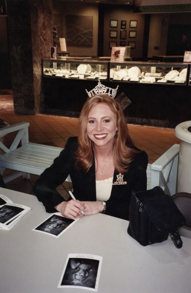 Miss Wisconsin 2002, Jayme Dawicki, wears a crown and sits at a table autographing photographs of herself.