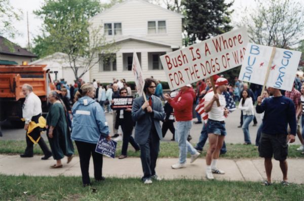 Protesters hold political signs outside Bush-Cheney rally. One sign reads: "Bush is a whore for the dogs of war" and the other reads: "Bush sucks."