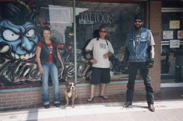 Two men, a woman, and a dog posing outdoors in front of the storefront of Mind Altering Tattoos.