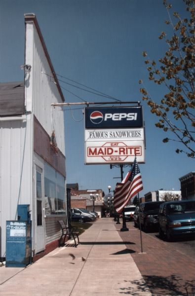 View down sidewalk of exterior of the Maid-Rite Restaurant, with hanging sign and American flag.