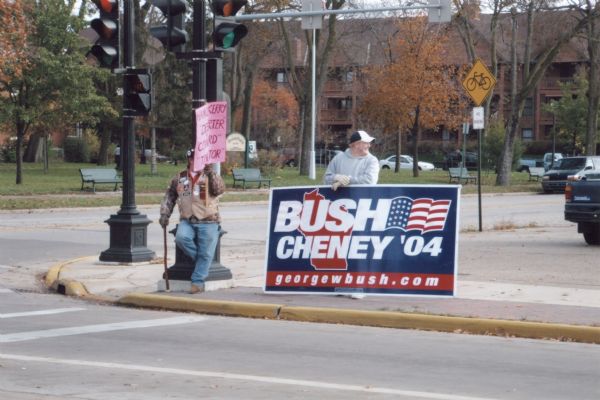 View from across street of a man holding Bush-Cheney '04 sign along intersection. A man next to him holds an illegible sign.