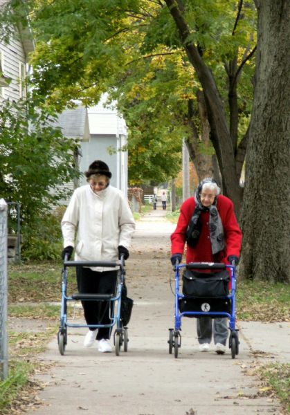 Two elderly women with walkers taking a stroll outdoors on a sidewalk.