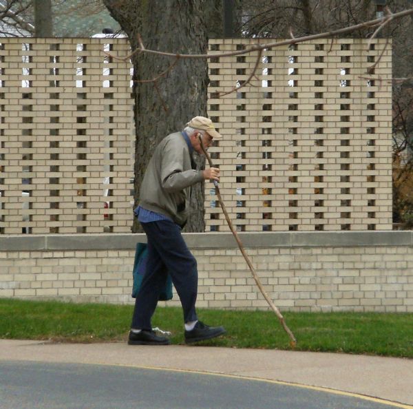 Elderly man walking outdoors while holding a bag and walking stick.