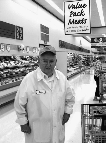 David A. Marcou, father of the photographer, and meat-cutter, stands underneath a sign in a store.