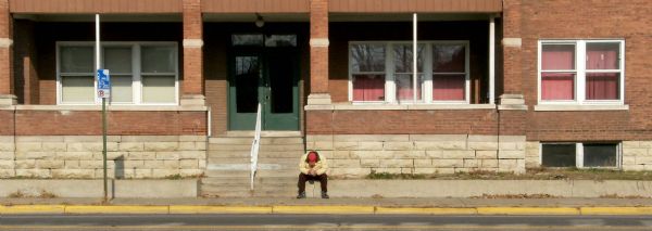 Panoramic view from across street of building near bus stop. A man is waiting for the bus near the steps to the building.