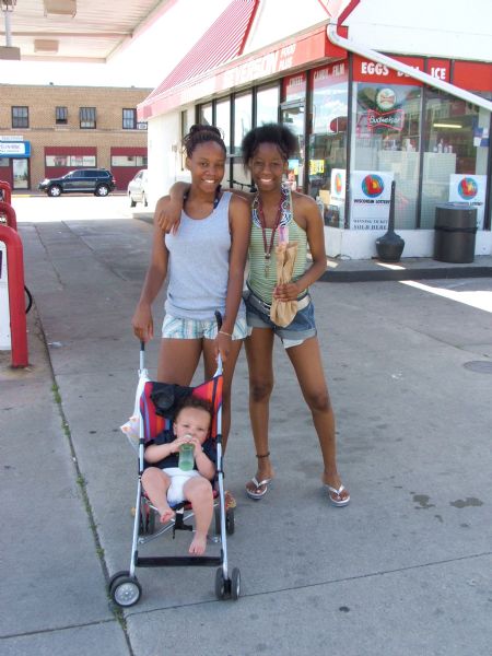 Two young women and an infant in a stroller posing for a picture at a gas station. One woma has her arm around the other woman.