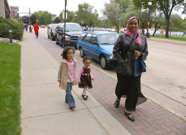 A woman is walking down the sidewalk, wearing a head covering and smiling at the camera, while two young girls are following close behind.