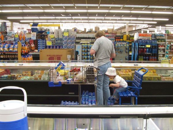 A child is sitting in a shopping cart while her mother is looking over frozen goods at a grocery store.