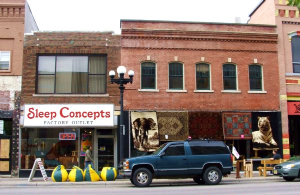 View from across street of exterior of "Sleep Concepts" store and rug store. Bean bags are set up on the sidewalk near a sandwich board sign.