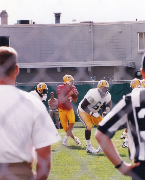 View through chain-link fence of Green Bay Packer Brett Favre, wearing a red scrimmage jersey, and other players at football practice.