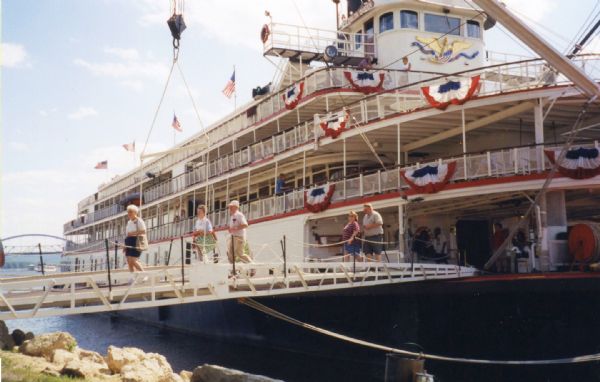 The "Delta Queen" steamboat at dock. People are walking across the gangplank toward the shoreline.
