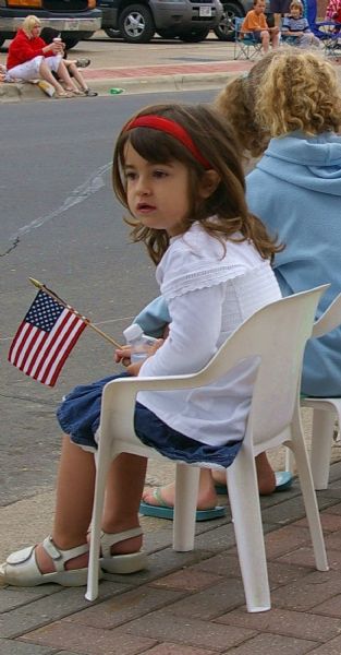 A young girl dressed in red, white, and blue is watching the Memorial Day parade while holding an American flag and sitting in a chair on the sidewalk.