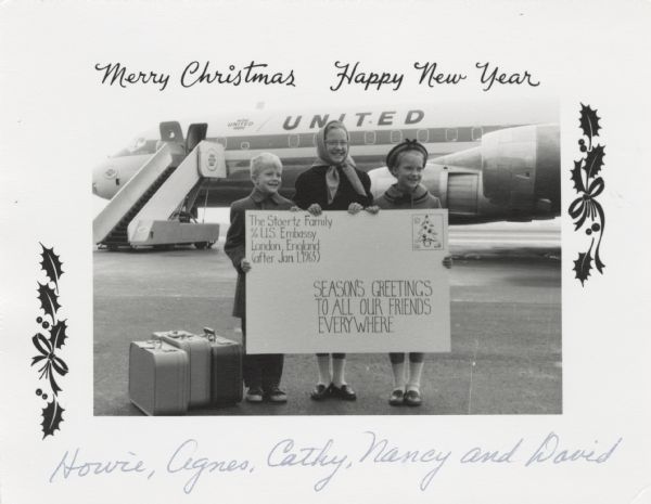 Photographic holiday greeting card of three children, one boy and two girls, dressed in winter clothes, holding up an enlarged postcard. On the postcard is the text: "The Stoertz Family, c/o U.S. Embassy, London, England, (after Jan. 1965), Season's Greetings To All Our Friends Everywhere." They are posed in front of an airplane at an airport. Their luggage is to their right. Holly decorates the sides of the postcard.