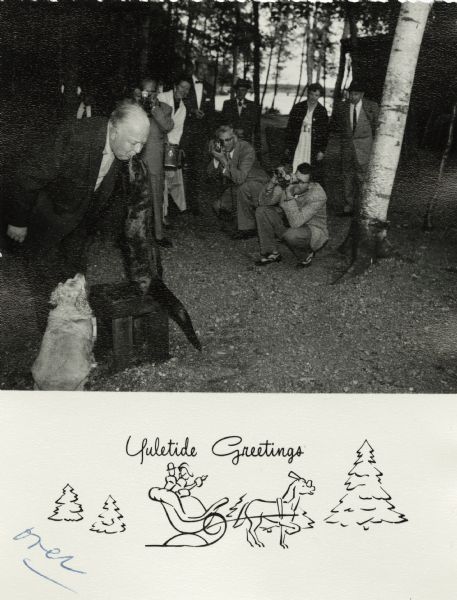 Photographic holiday card featuring Carl Marty of the Northernaire Resort in Three Lakes, Wisconsin. He is famous for his hospitality and his "animal friends." Here he is shown with Sugar the Otter, and Ginger his Cocker Spaniel. He is being photographed by visitors. The text "Yuletide Greetings" appears below with a drawing of a couple in a horse-drawn sleigh.