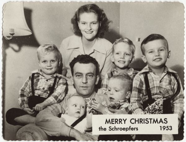 Photographic holiday card of the Schroepfer family. They are gathered around the father seated in an armchair. The mother stands behind the chair. Four brothers are wearing matching plaid shirts. The father is holding an infant. In the lower right corner it says "Merry Christmas, The Schroepfers 1953."