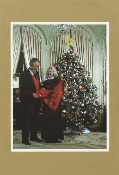 Photographic Christmas card from Governor and Mrs. Lee Sherman Dreyfus, governor of Wisconsin 1979-1983. Front of card depicts the governor and his wife in front of their Christmas tree. The image has a white border on a metallic gold background. Mrs. Dreyfus is showing her husband an embroidered red Christmas vest. The inside of the card reads: "He has given us a 23rd Psalm place in which to live; He has given us life and all the marvelous senses to savor it; He has given us minds with unlimited capacity to imagine and create, and He has given us a Saviour (misspelled in card), His own beloved son. How He must love us. Lee Sherman Dreyfus".